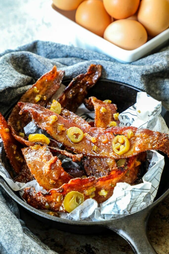 Bacon with jalapeños on top in a black skillet with eggs in the background.