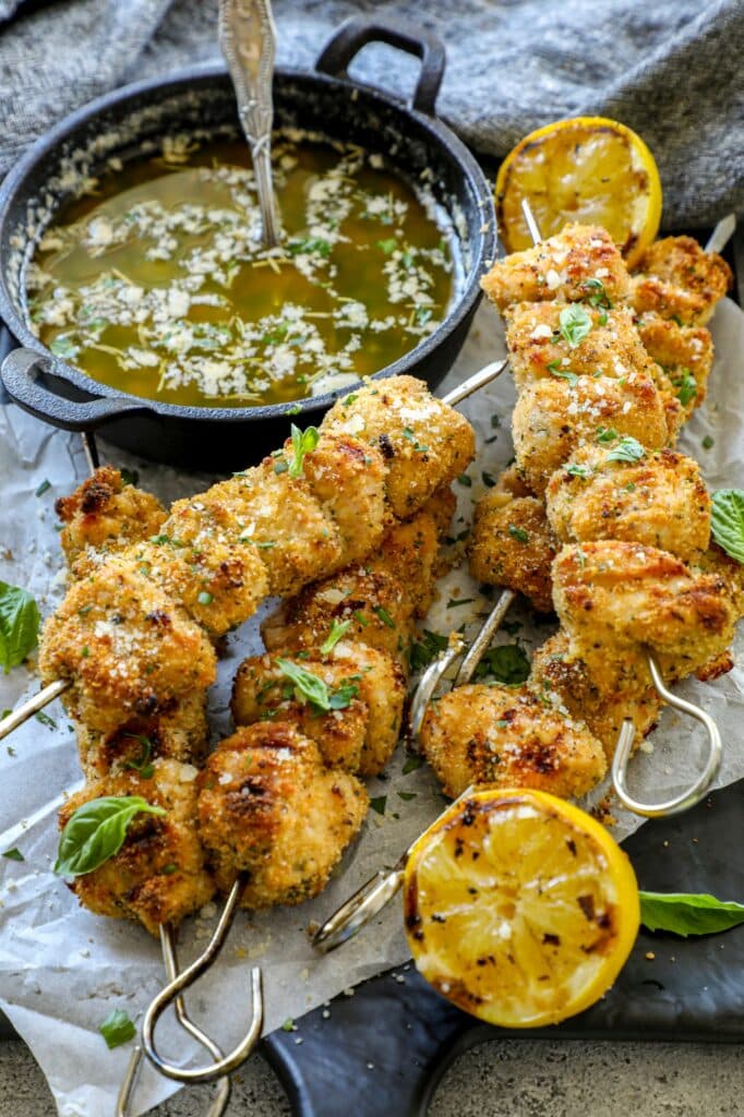 Breaded chicken skewers stacked on top of each other next to a cast iron pan with sauce in it.