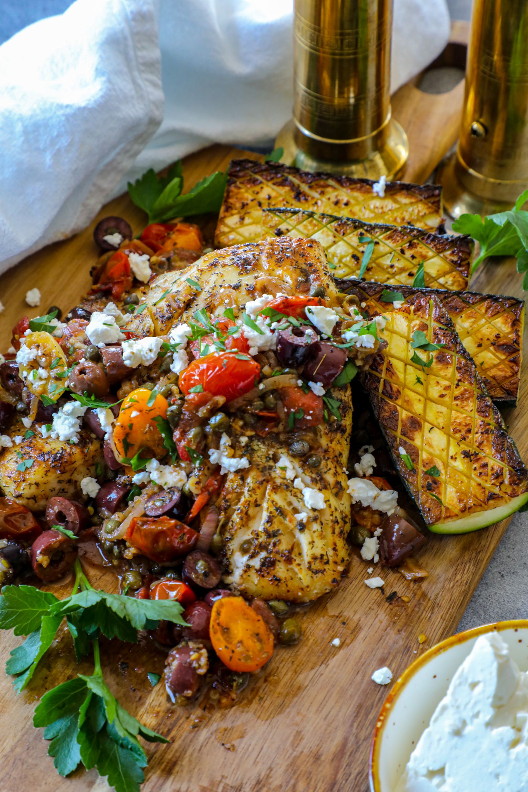 Cod on a wooden cutting board with veggie mixture on top and grilled zucchini on the side.